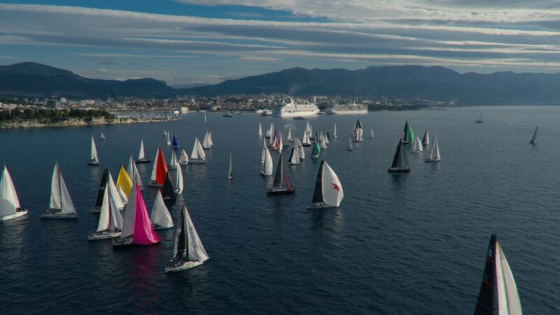 A fleet of sailboats with white, pink, yellow and red sails on a bay, with a city and mountains behind.