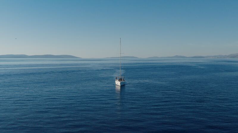 A single sailboat on open, calm sea with islands on the horizon.