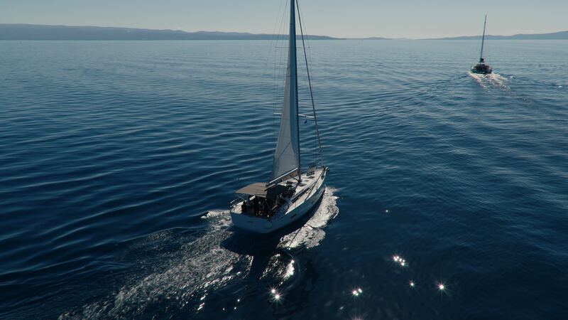 A sailboat under main and headsail crosses rippled blue water, seen from above and behind, with a second boat ahead.